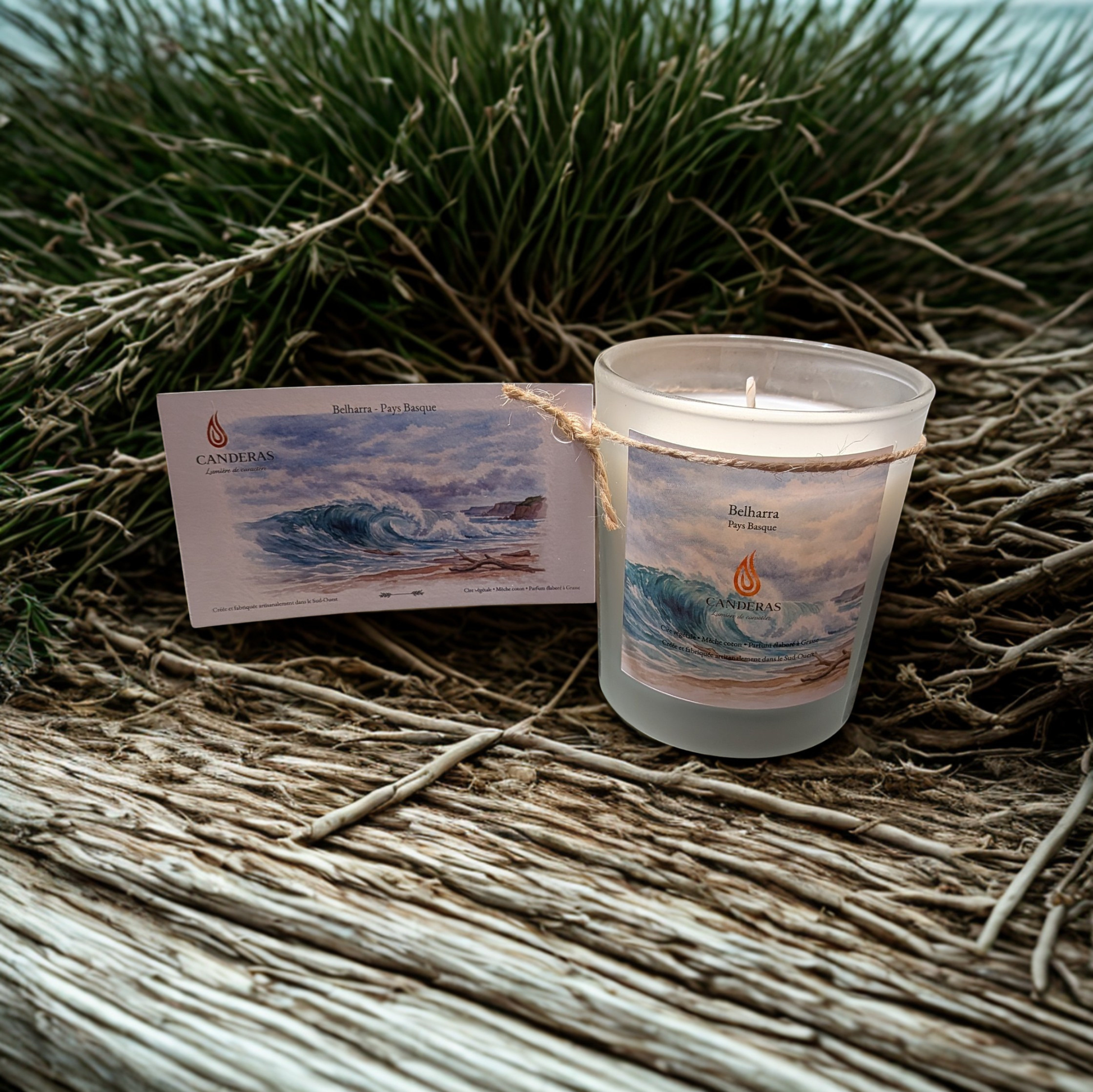 Bougie artisanale du Sud-Ouest Candèras Belharra avec carte postale assortie sur fond bois clair, textile naturel et mur texturé.